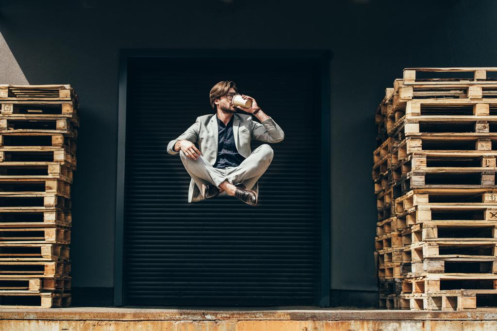 knappe jonge zakenman zwevend in de lucht in lotus houding en drinken koffie op industriële locatie voor roller poort  - Foto, afbeelding