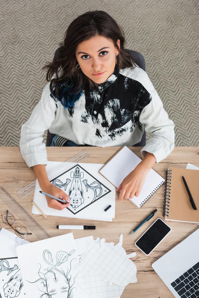 vista aérea da pintura artista feminina e olhando para a câmera enquanto sentado à mesa com laptop e smartphone
  - Foto, Imagem