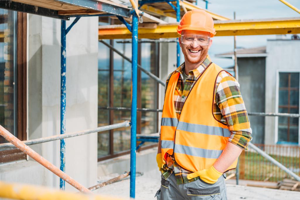 construtor sorrindo bonito em colete reflexivo e chapéu duro olhando para a câmera no local de construção
 - Foto, Imagem