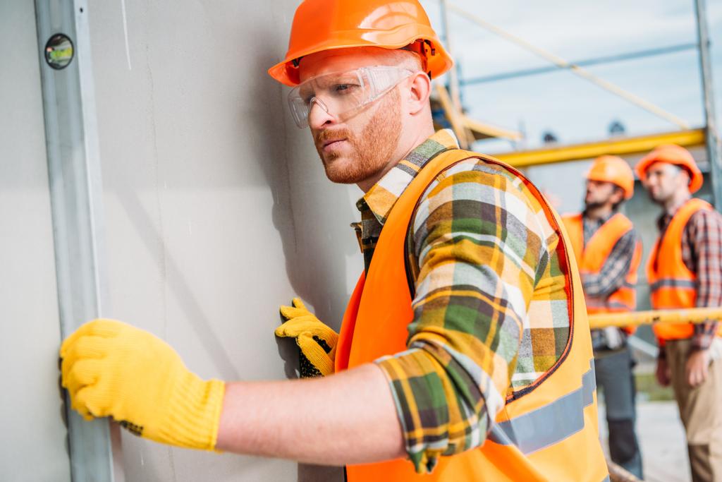 konzentrierter Bauarbeiter mit Blasenhöhe auf der Baustelle, während seine Kollegen im Hintergrund stehen - Foto, Bild
