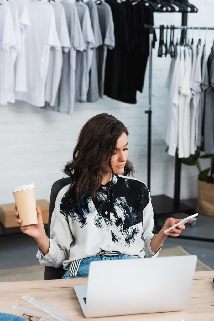 aantrekkelijke vrouwelijke modeontwerper met koffie beker met behulp van smartphone aan tafel met laptop  - Foto, afbeelding