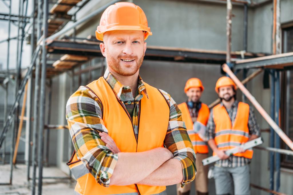 lachende bouwer permanent op de bouwplaats met gekruiste armen terwijl zijn collega's permanent op de achtergrond wazig - Foto, afbeelding