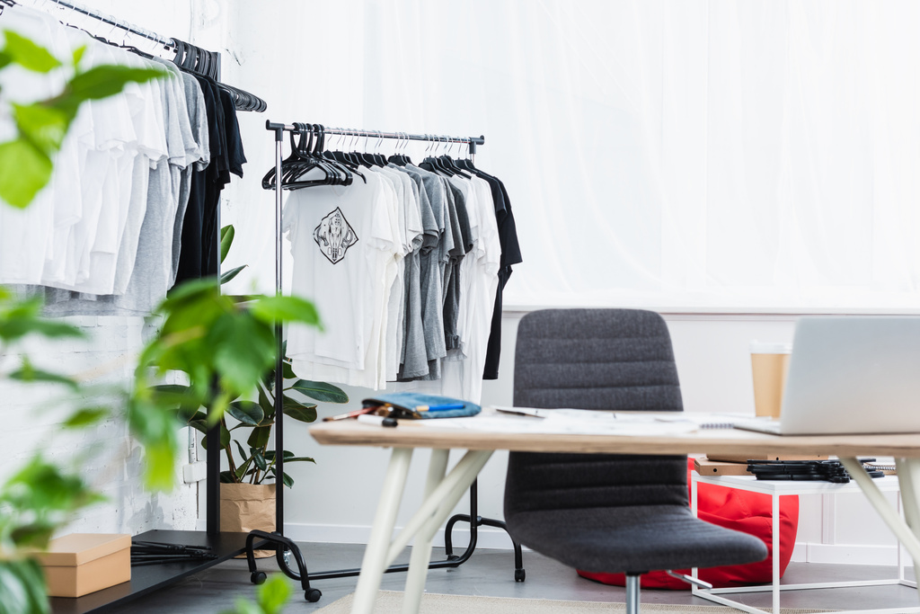 selectieve aandacht voor t-shirts op hangers en werktafel met laptop in kleding ontwerpstudio - Foto, afbeelding