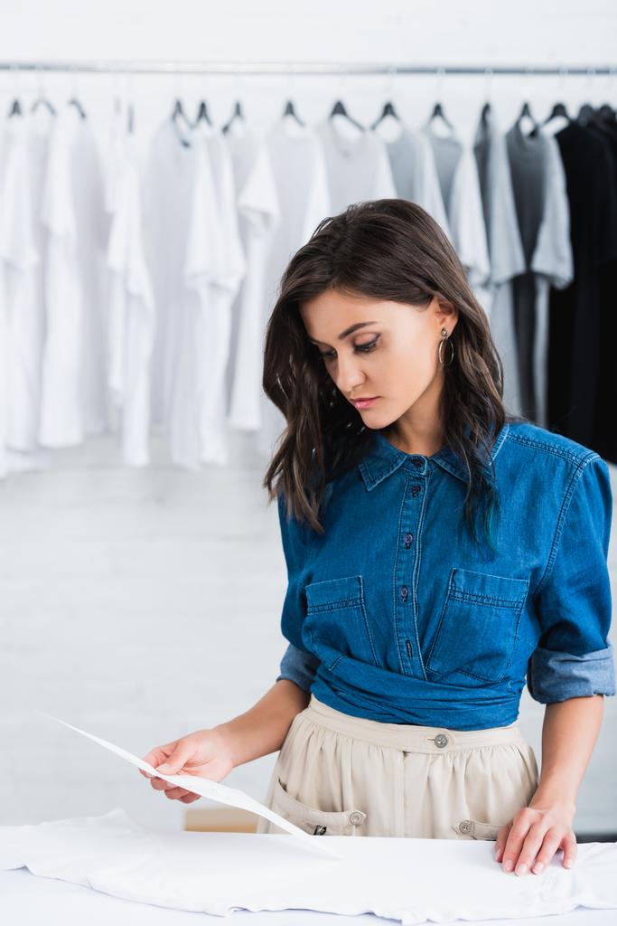 joven y atractiva diseñadora de moda femenina eligiendo la impresión para la camiseta blanca vacía en el estudio de diseño de ropa
 - Foto, Imagen