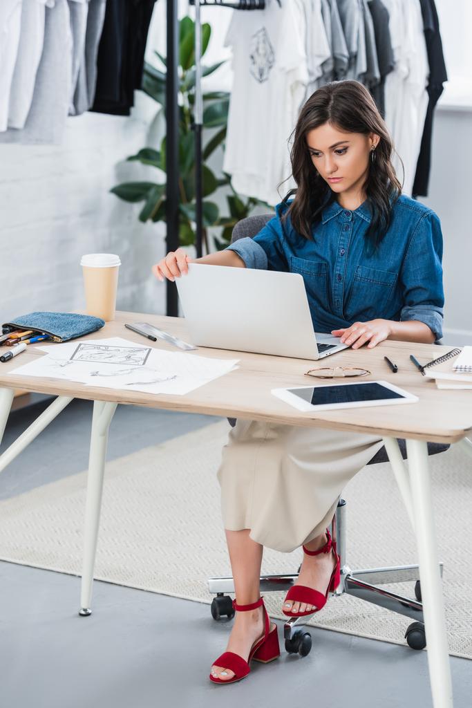 серьезная женщина модельер работает за столом с ноутбуком в студии
 - Фото, изображение