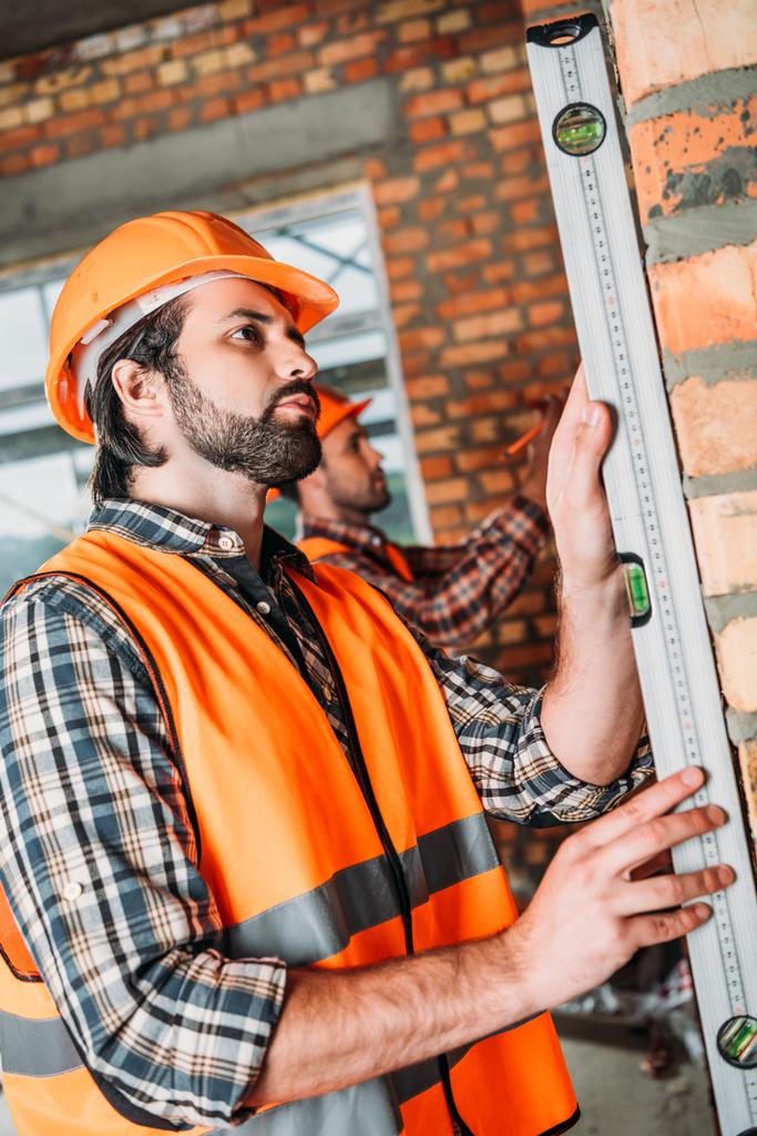 vista lateral do construtor bonito usando nível de bolha no local de construção, enquanto seu colega trabalhando em segundo plano
 - Foto, Imagem