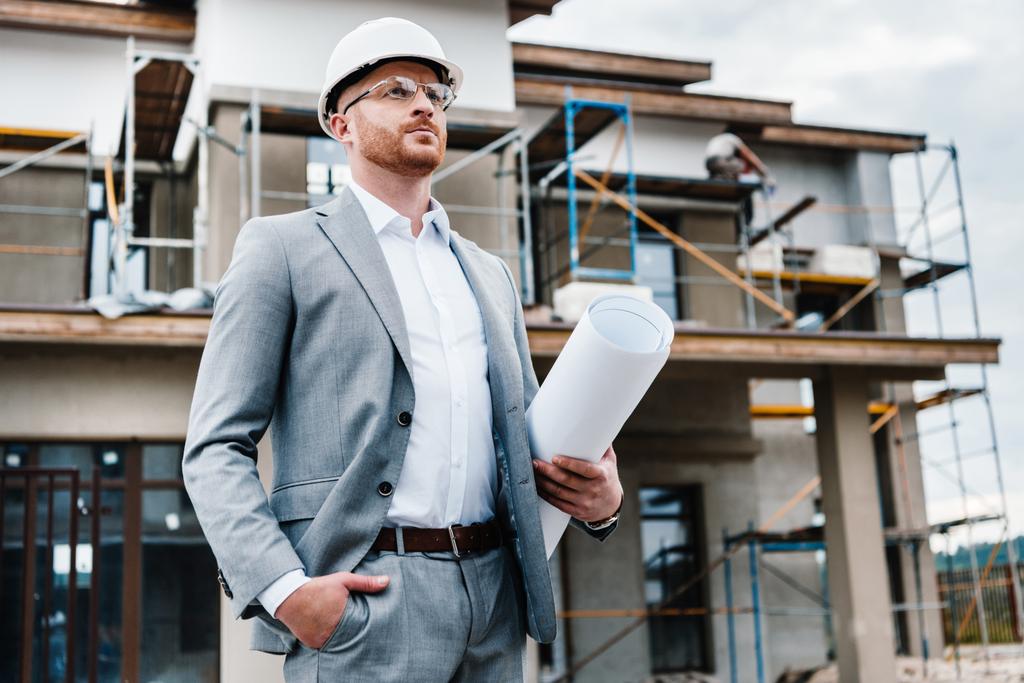 handsome architect in suit and hard hat holding blueprint standing in front of building house and looking away - Photo, Image