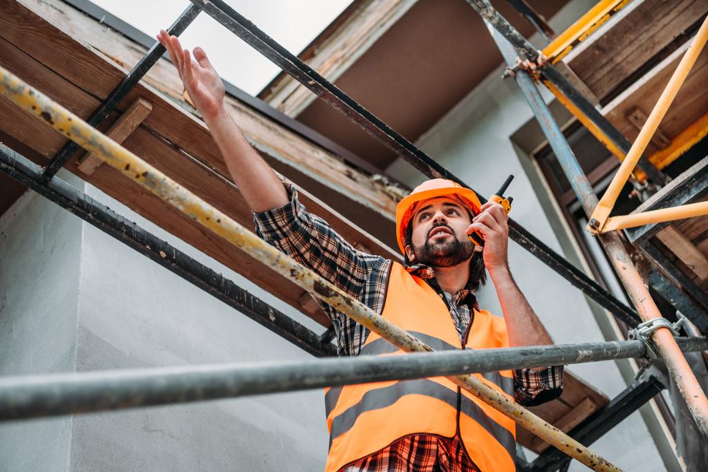 Onderaanzicht voor builder in beschermende helm en vest met walkie talkie op bouwplaats - Foto, afbeelding