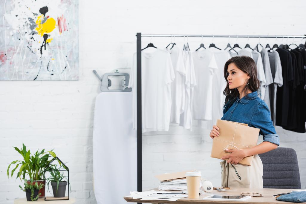 atraente feminino designer segurando pacote de papel no estúdio de design de roupas
 - Foto, Imagem
