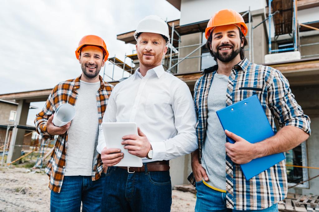 group of smiling architects in hard hats looking at camera in front of building house - Photo, Image