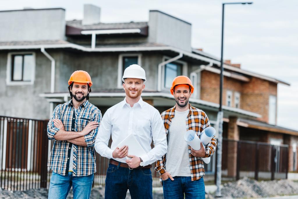 group of smiling architects in hard hats looking at camera on building street - Photo, Image