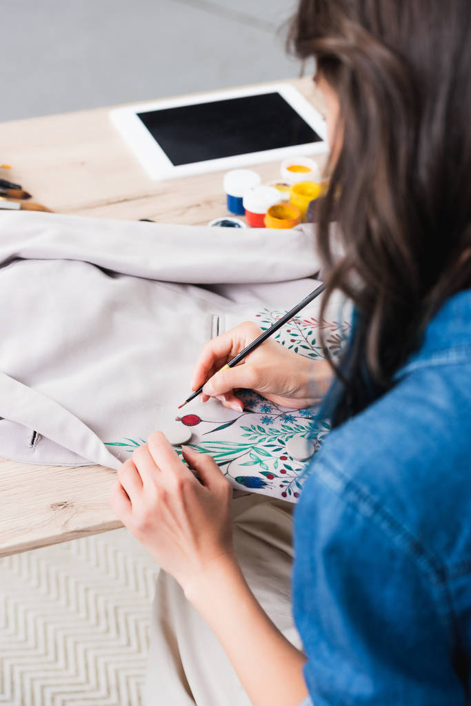 vrouwelijke mode-ontwerpster schilderen op jas op werktafel in kleding ontwerpstudio - Foto, afbeelding