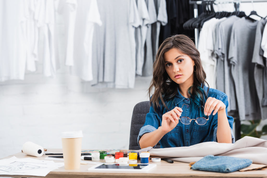 junge Modedesignerin sitzt mit Farben und Kaffee am Arbeitstisch - Foto, Bild