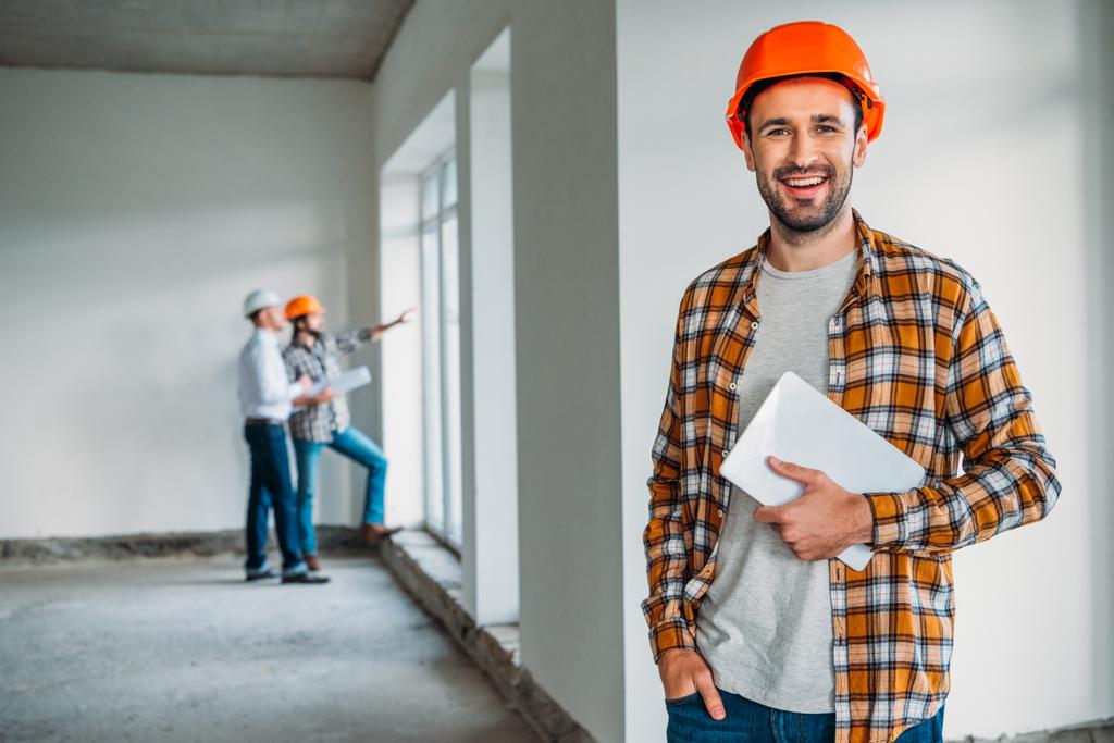 bel architecte en chemise à carreaux et chapeau dur debout à l'intérieur de la maison de construction avec tablette dans les mains
 - Photo, image