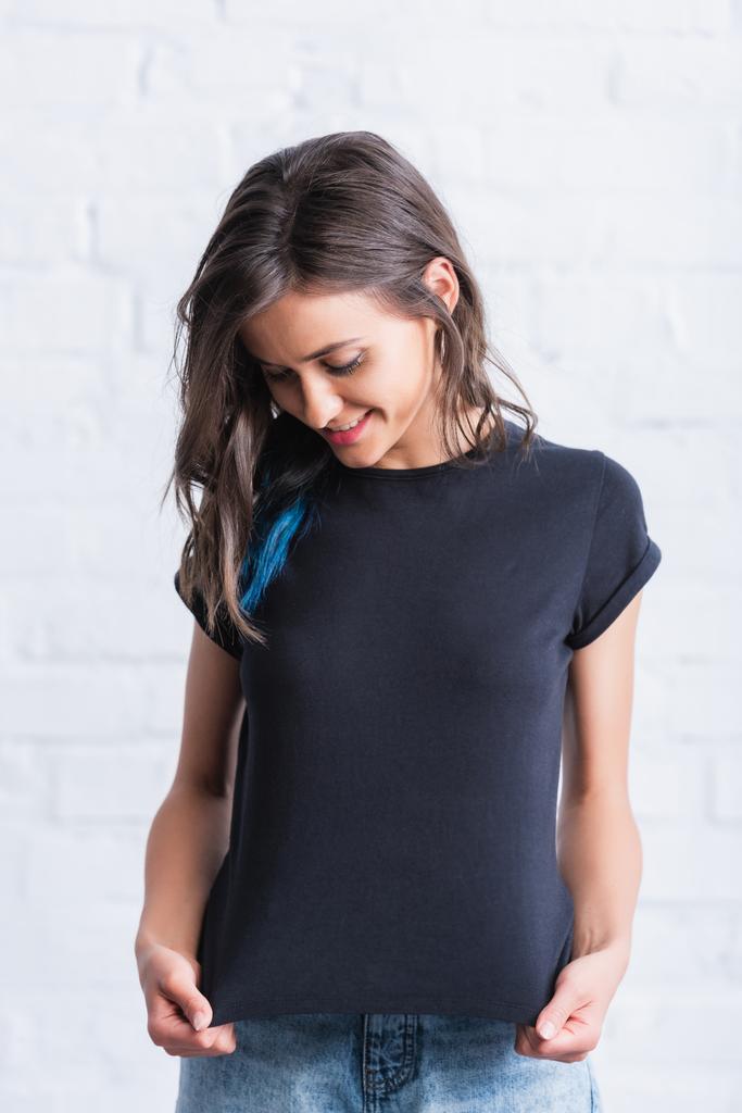 Lächelnde junge Frau zeigt leeres schwarzes T-Shirt vor Backsteinmauer  - Foto, Bild