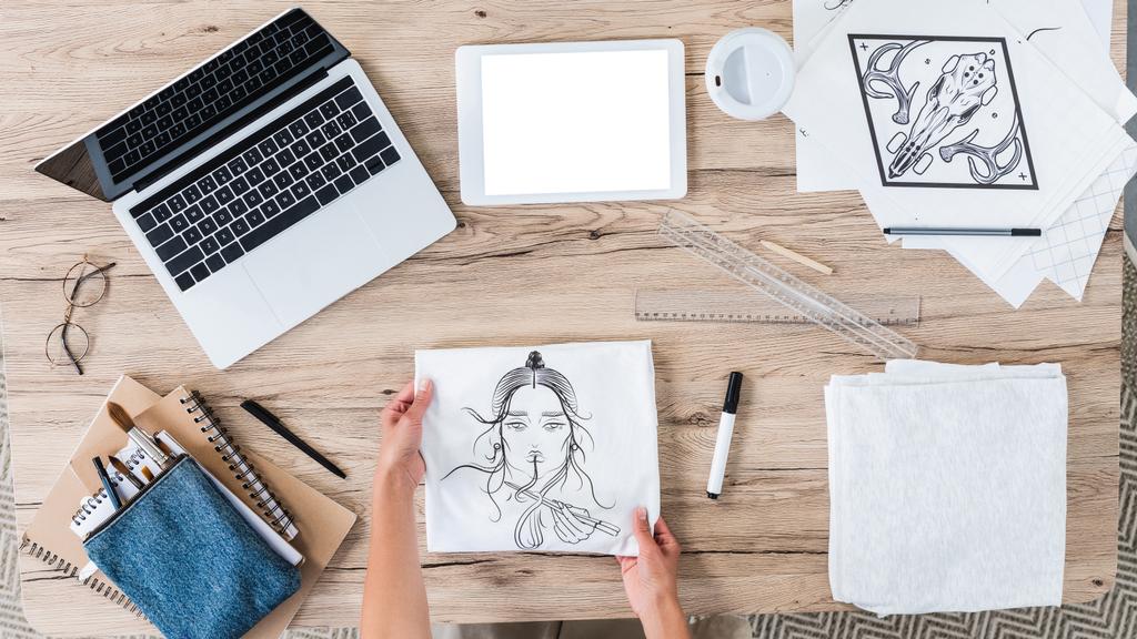 vista aérea do designer feminino colocando t-shirt branca com impressão na mesa com dispositivos digitais uma pintura
  - Foto, Imagem