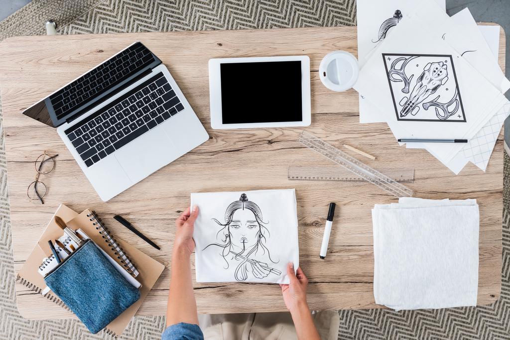cropped image of female designer putting white t-shirt with print on table with digital devices an paintings  - Photo, Image