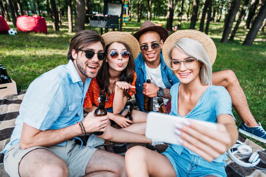 smiling multicultural friends with beer taking selfie on smartphone while resting in summer park - Photo, Image
