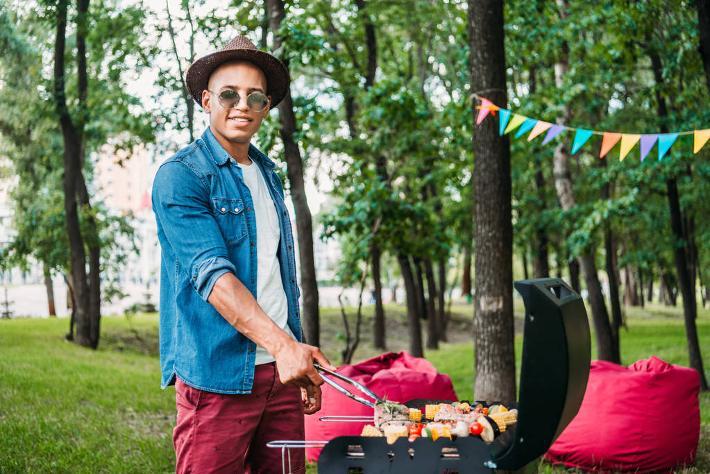 Afro-Amerikan erkek güneş gözlüğü ve şapka yemek yemek ızgara Park yan görünüm - Fotoğraf, Görsel