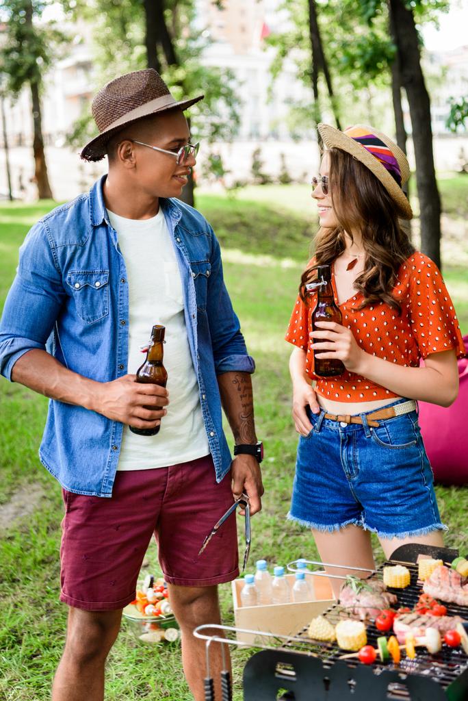 χαμογελώντας Διαφυλετικό ζευγάρι με μπύρα στέκεται κοντά στο γκριλ στο πάρκο καλοκαίρι - Φωτογραφία, εικόνα