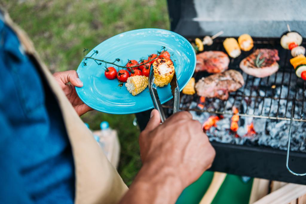 vue partielle de l'homme afro-américain wit tongs mettre des légumes grillés sur l'assiette pendant le barbecue dans le parc
 - Photo, image