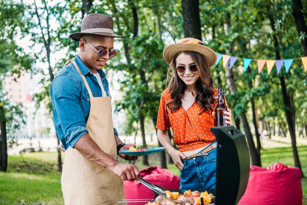 donna sorridente con birra guardando il fidanzato afro-americano che cucina cibo sulla griglia durante il barbecue
 - Foto, immagini