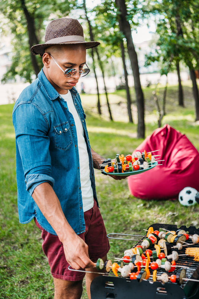 вид сбоку на африканского американца в солнцезащитных очках, принимающего на гриле овощи во время барбекю в парке
 - Фото, изображение