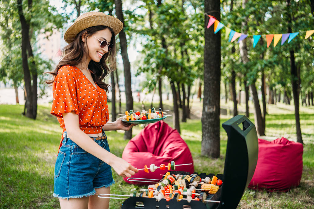 вид сбоку улыбающейся молодой женщины, которая берет овощи из гриля в парке
 - Фото, изображение