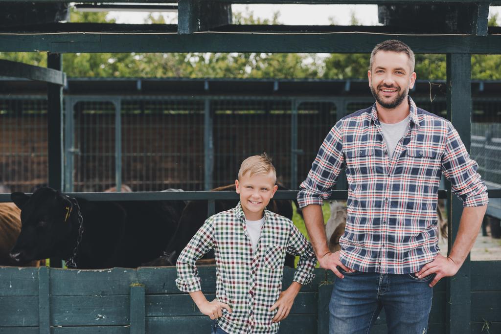 felice padre e figlio in piedi con le mani in vita e sorridente alla fotocamera in fattoria
 - Foto, immagini