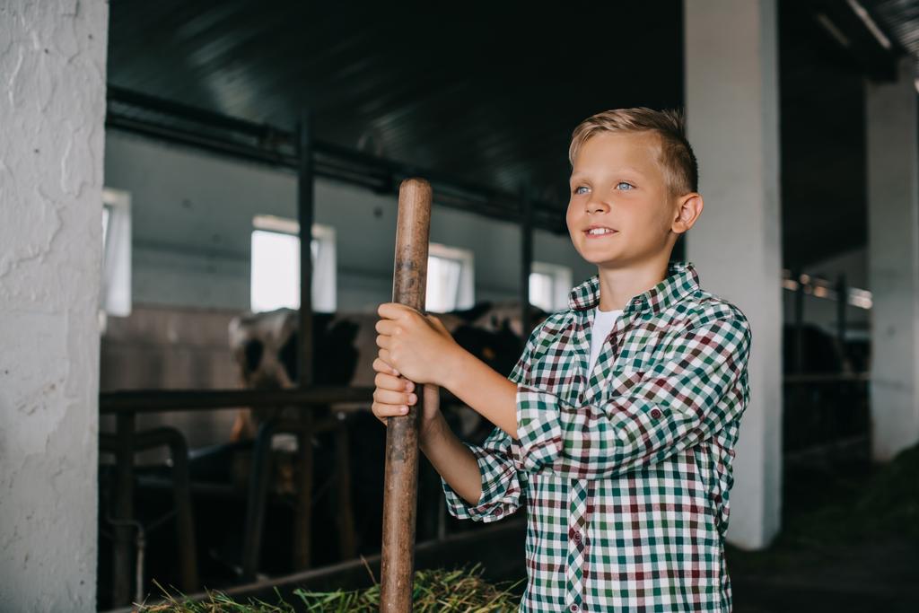 smiling boy holding rake and looking away while working at cowshed - Photo, Image