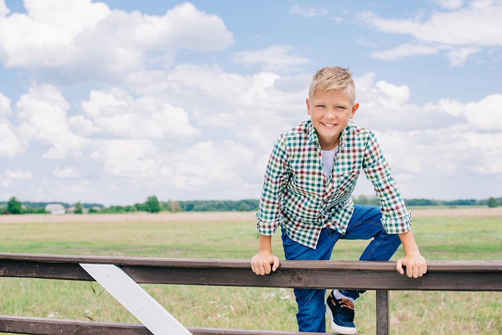 happy boy climbing on fence and smiling at camera on farm - Photo, Image