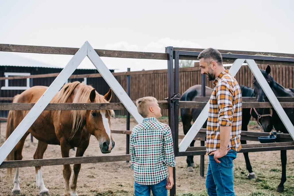 Widok z ojcem i synem uśmiechający się sobą stojąc w pobliżu konie na ranczo tyłu - Zdjęcie, obraz