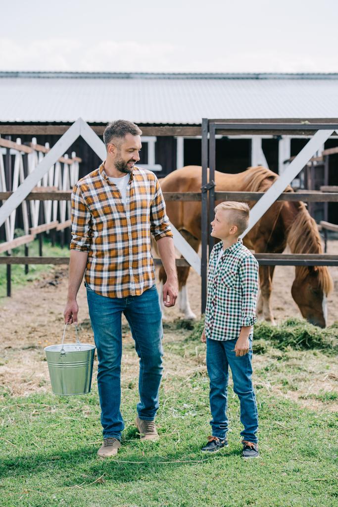 otec s kbelík a malý syn usmívající se navzájem stoje u koní na ranči - Fotografie, Obrázek