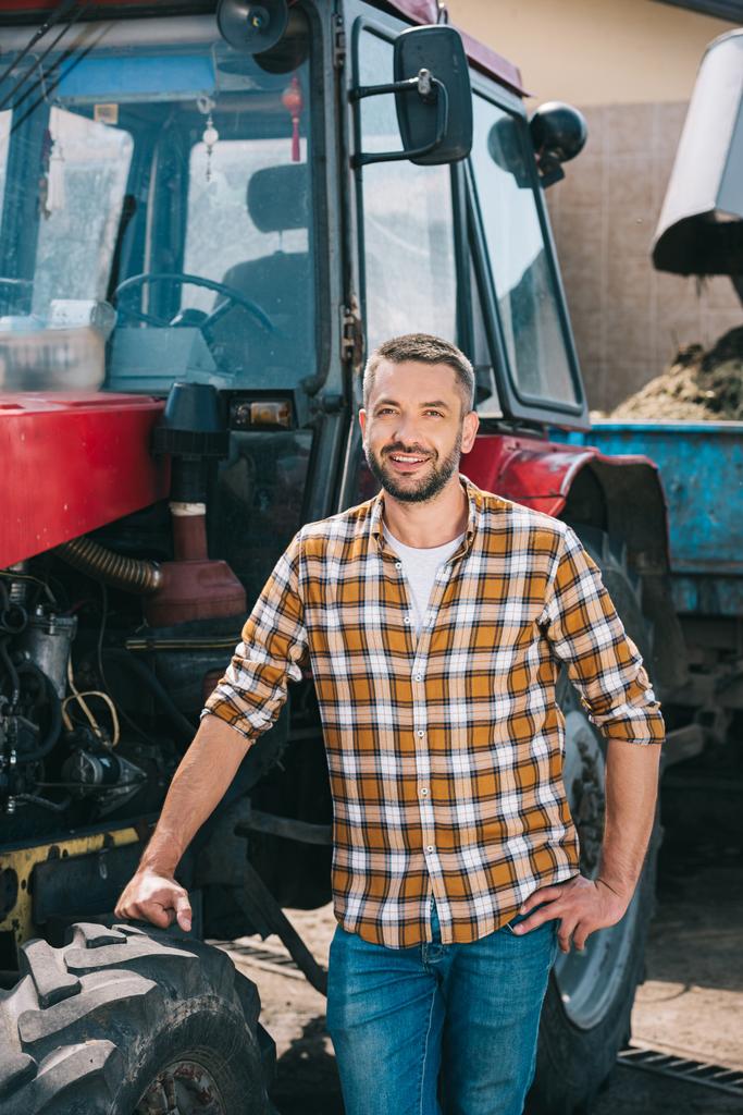 handsome middle aged farmer standing near tractor and smiling at camera - Photo, Image