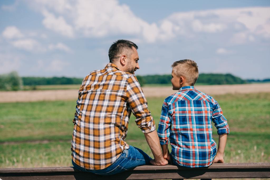 Achteraanzicht van vader en zoon zittend op het hek en glimlachen elkaar op boerderij - Foto, afbeelding
