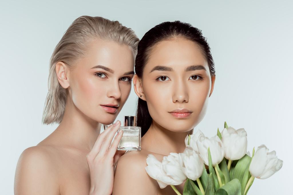 attractive multicultural girls posing with tulips and bottle of perfume, isolated on grey - Photo, Image
