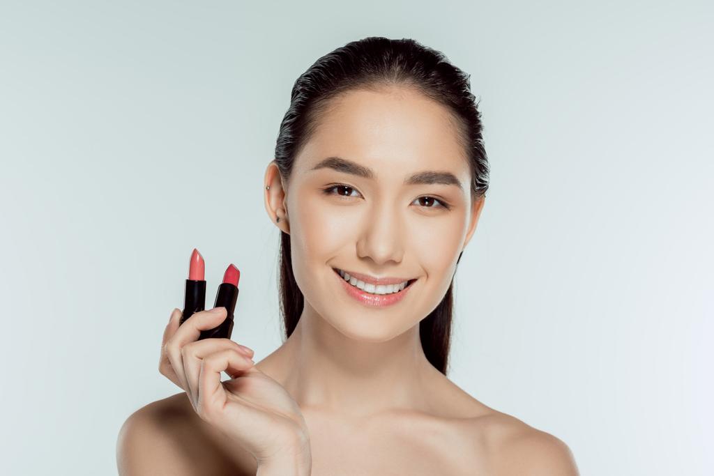 attraktive lächelnde asiatische Frau mit zwei Lippenstiften, isoliert auf grau - Foto, Bild