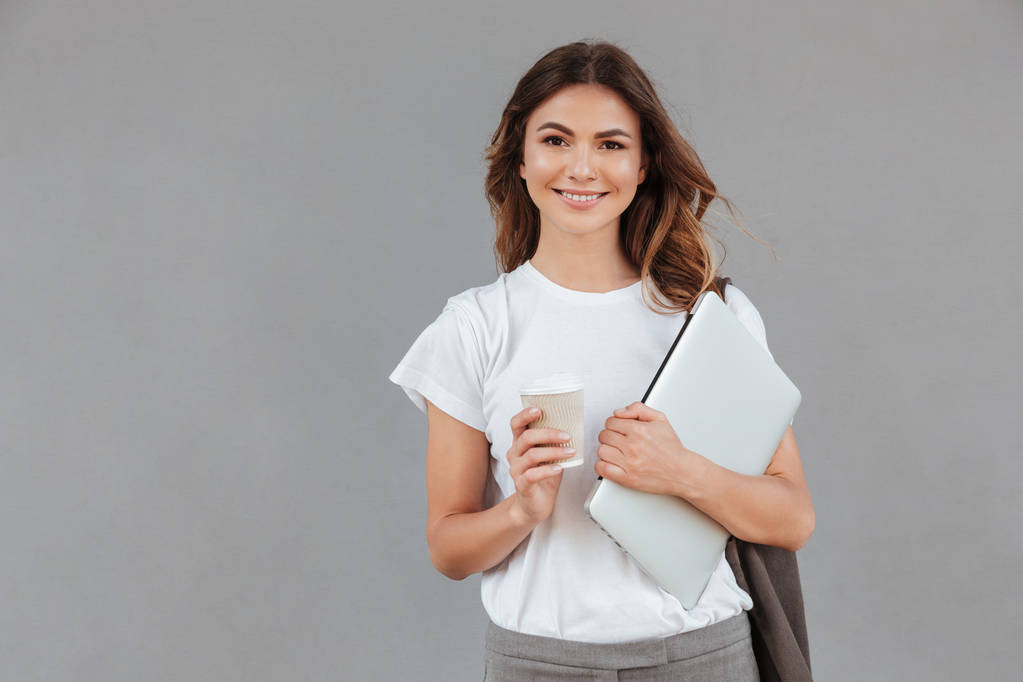 Фото красивої молодої жінки, яка посміхається і стоїть на сірому фоні ізольовано срібним ноутбуком і виймає каву в руках
 - Фото, зображення