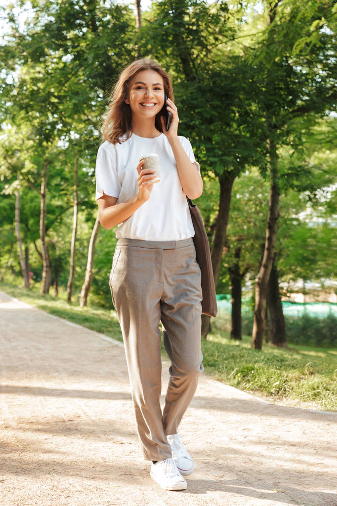 Bild der schönen brünetten Frau, die an einem Sommertag einen schmalen Pfad durch den grünen Park hinuntergeht und mit einer Tasse Kaffee zum Mitnehmen auf dem Smartphone spricht - Foto, Bild