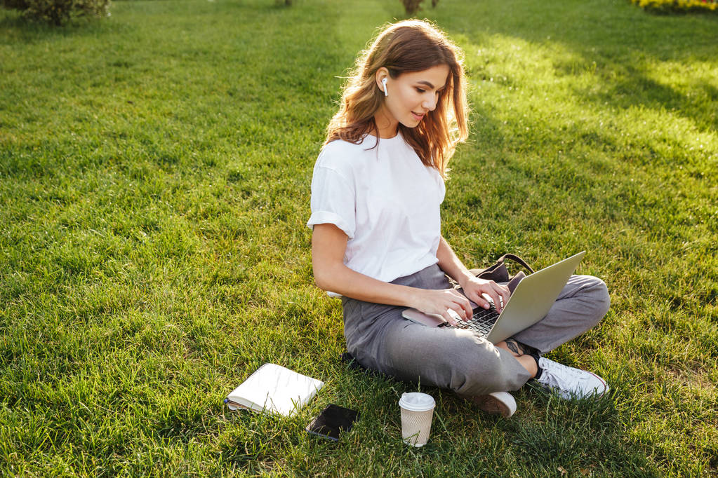 Foto da mulher europeia 20s sentado na grama verde no parque com pernas cruzadas durante o dia de verão, enquanto usando laptop e fone de ouvido bluetooth
 - Foto, Imagem