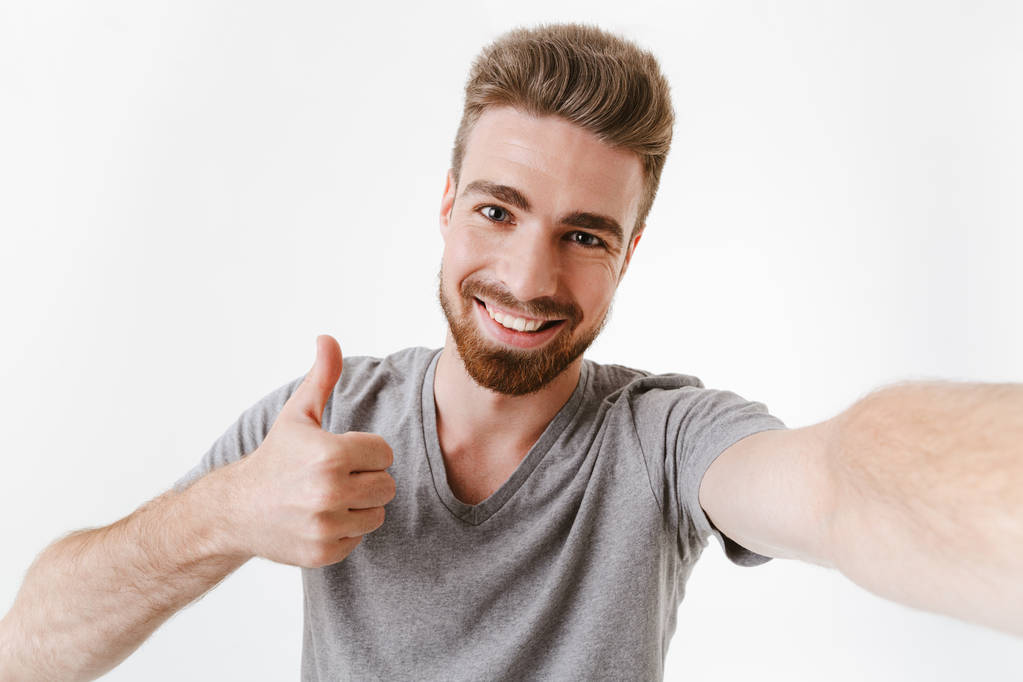 Beeld van knappe tevreden jonge man boven witte muur achtergrond op zoek camera geïsoleerd staan duimschroef opwaarts gebaar maken een selfie nemen. - Foto, afbeelding