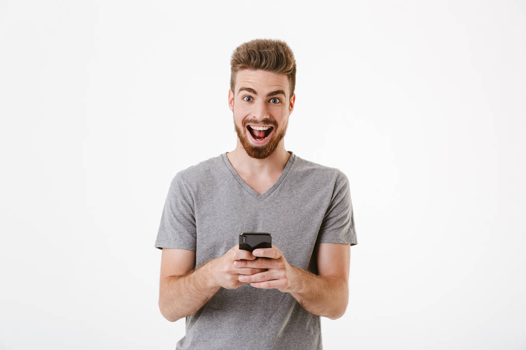 Bild eines glücklichen, gut aussehenden jungen Mannes, der isoliert über weißem Wandhintergrund mit dem Handy steht. Kamera gesucht. - Foto, Bild