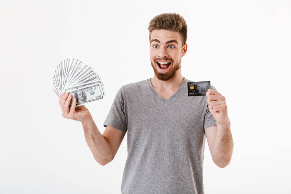 Para ve kredi kartı tutan beyaz duvar arka plan üzerinde izole ayakta heyecanlı mutlu genç adam görüntüsünü. - Fotoğraf, Görsel