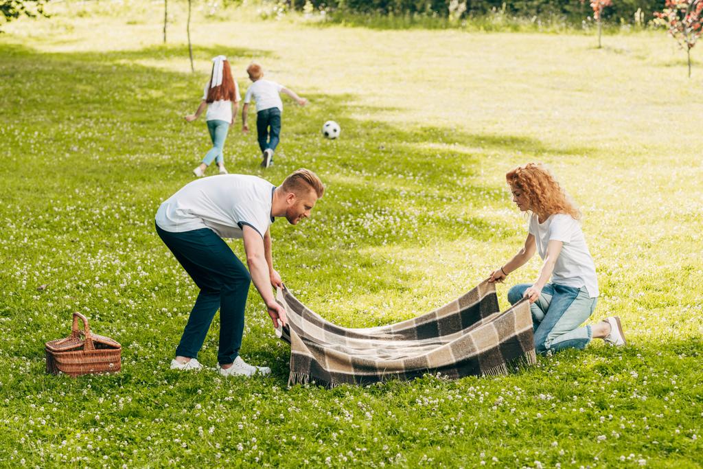 parents holding plaid while kids playing with ball at picnic in park - Photo, Image