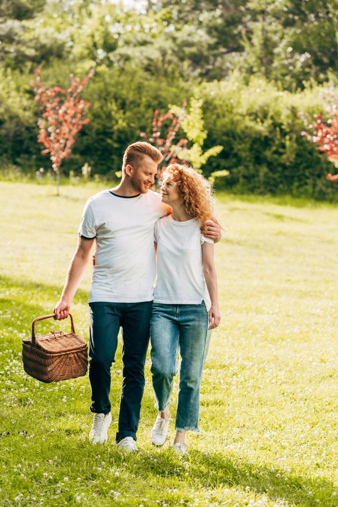 glückliches junges Paar mit Picknickkorb spaziert auf Rasen im Park - Foto, Bild