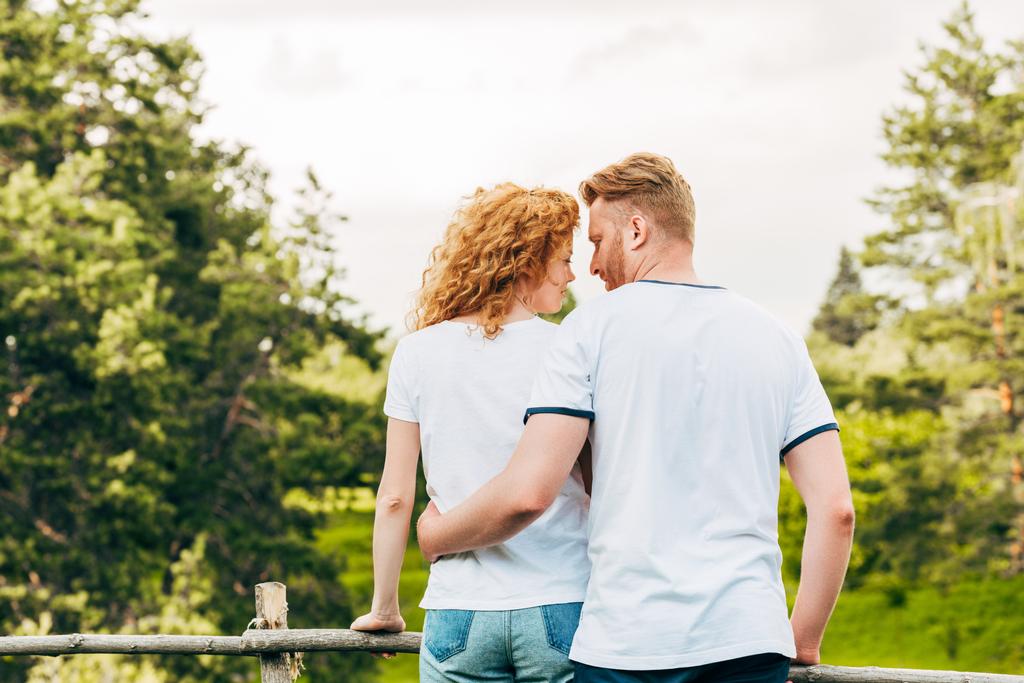 arkadan görünüşü kucaklayan ve birbirlerine birlikte parkta ayakta iken gülümseyen bir kızıl saçlı çift - Fotoğraf, Görsel