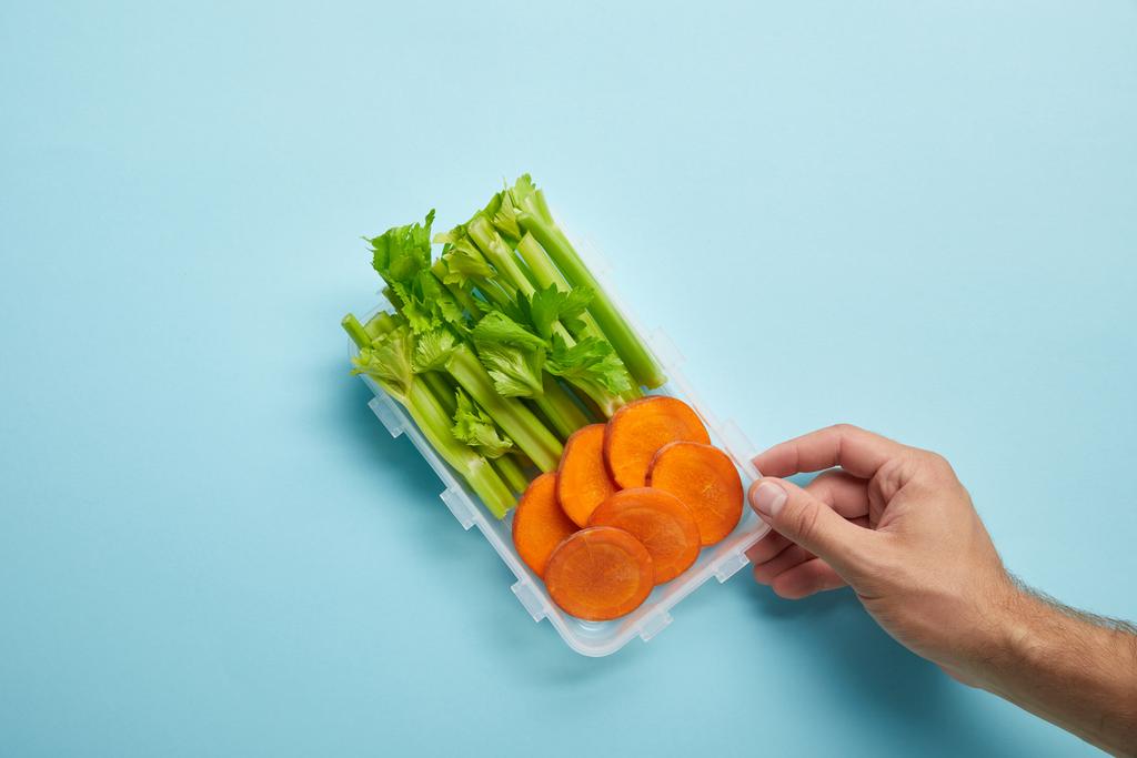 обрізаний знімок чоловічої руки та харчовий контейнер, повний селери та шматочків моркви ізольовані на синьому
 - Фото, зображення