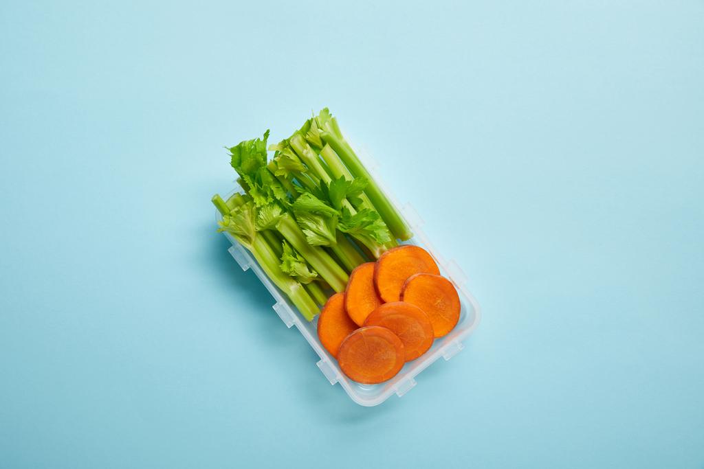 vista dall'alto del contenitore alimentare pieno di sano sedano e fette di carota isolate su blu
 - Foto, immagini