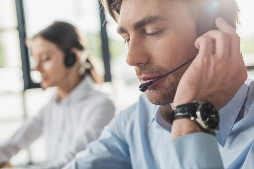bonito call center gerente em fones de ouvido com microfone sentado no local de trabalho, enquanto seu colega sentado em segundo plano
 - Foto, Imagem