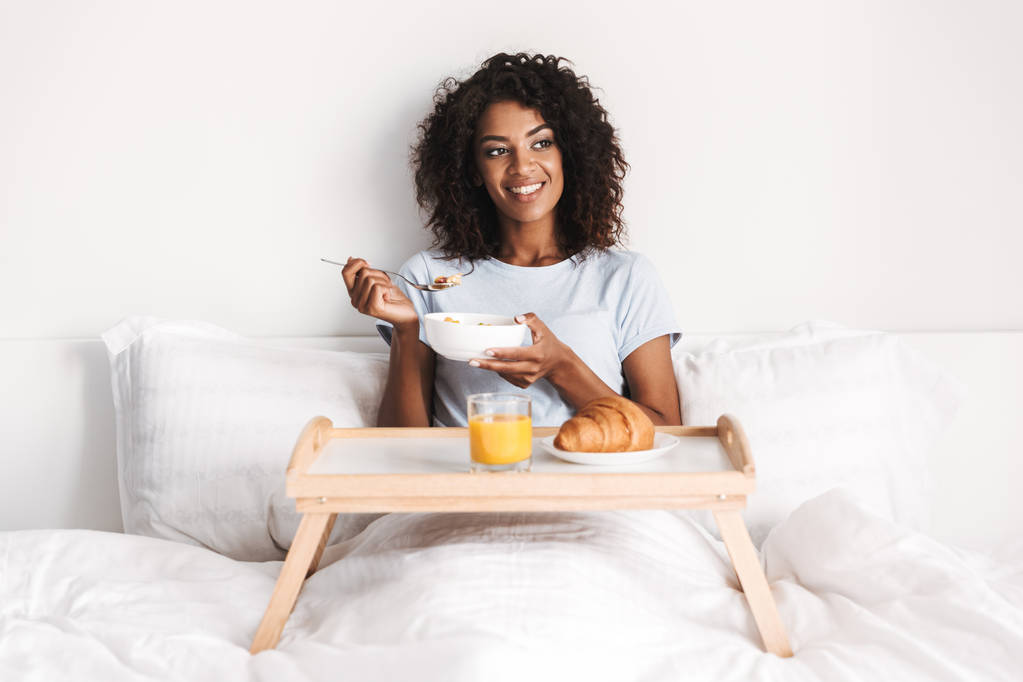 Mosolygó fiatal afrikai nő, ízletes reggeli elfogyasztására a tálcán az ágyban - Fotó, kép